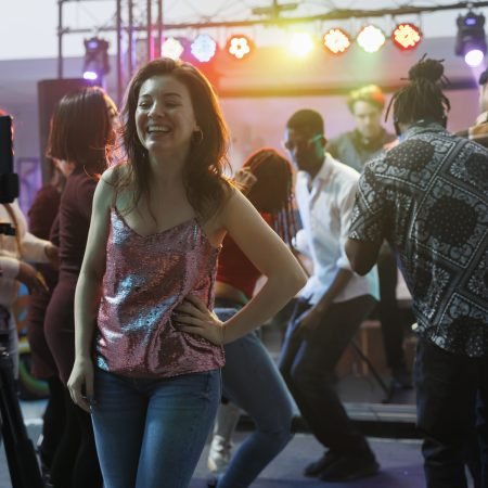Cheerful smiling woman taking photo on smartphone while having fun in nightclub. Carefree laughing girl recording video using mobile phone on tripod while partying in club